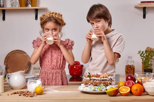 Little good-looking boy dressed in a light t-shirt and jeans and a pretty girl wearing in a pink dress are making a cake at a kitchen, against a white wall with shelves on it. They are drinking milk and looking away.