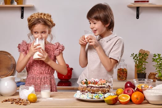 Little pretty kid dressed in a light t-shirt and jeans and a beautiful girl wearing in a pink dress are making a cake at a kitchen, against a white wall with shelves on it. They are drinking milk and smiling.