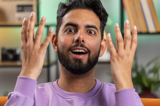 Oh my God, Wow. Young indian man surprised looking at camera with big eyes, shocked by sudden victory, good win news, celebrating. Portrait of excited amazed hindu guy at home apartment room on sofa