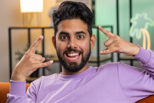 Overjoyed delighted young indian man showing rock n roll gesture by hands, cool sign, shouting yeah with crazy expression, dancing. Hindu guy emotionally rejoicing in success at home living room couch