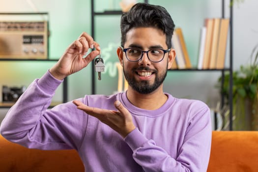 Happy young indian man real estate agent lifting hand showing the keys of new home house apartment, buying or renting property, mortgage loan. Portrait of hindu guy at home apartment room on sofa
