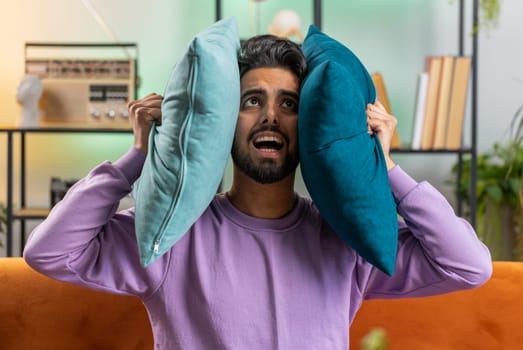 Repair work at neighbours. Irritated indian man relaxing on couch cover ears with pillows annoyed by noisy neighbors suffer from headache wish silence. Thin walls at home flat without sound insulation