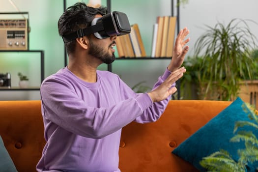Indian man using virtual reality futuristic technology VR app headset helmet to play simulation 3D 360 video game, watching film movie at modern home apartment. Hindu guy in goggles sitting on couch