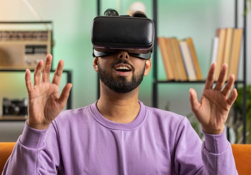 Indian man using virtual reality futuristic technology VR app headset helmet to play simulation 3D 360 video game, watching film movie at modern home apartment. Hindu guy in goggles sitting on couch