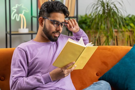 Young indian man reading interesting book, turning pages smiling enjoying literature, taking a rest on comfortable couch. Portrait of peaceful cheerful hindu guy at home apartment living room on sofa