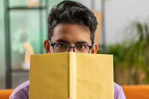 Young indian man in glasses reading interesting book, hiding behind book, enjoying literature, taking a rest on couch. Portrait of peaceful cheerful hindu guy at home apartment living room on sofa