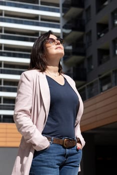 Relaxed Mature Female With Closed Eyes Enjoys Sun, Peace In Apartment Campus, Complex In Daytime. Female Brunette Wears Casual Clot. Modern Residential Building, Vertical Plane. High quality photo