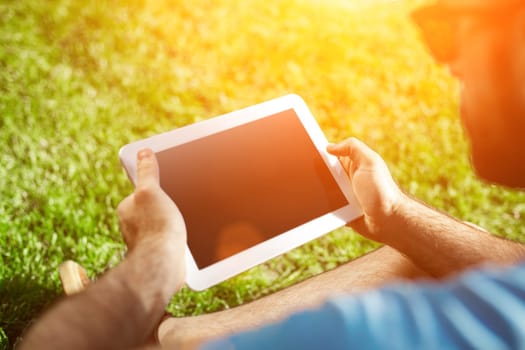 Young man using and typing tablet computer in summer grass. Freelancer working in outdoor park. Sun flare