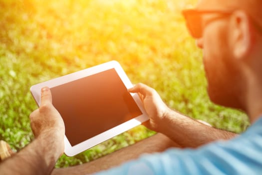 Young man using and typing tablet computer in summer grass. Freelancer working in outdoor park. Sun flare