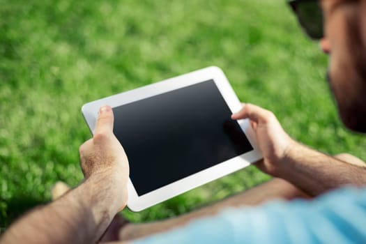 Young man using and typing tablet computer in summer grass. Freelancer working in outdoor park