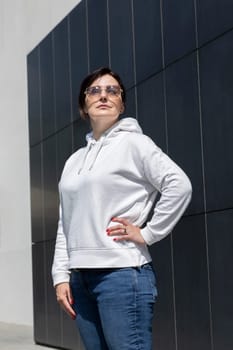 Closeup Pretty Mature Brunette Woman in White Hoody, Jeans and Sunglasses Looks Away On Dark Background Wall. Sportive Confident 40 Yo Beautiful White Woman in Urban Area Vertical Plane. Lifestyle.