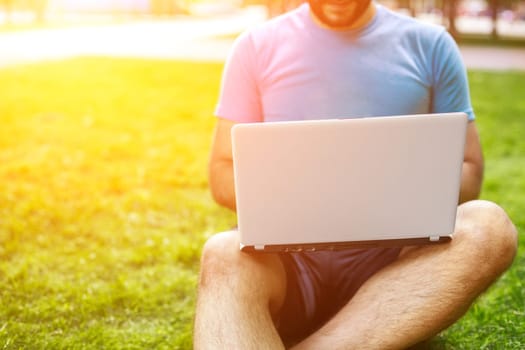 Young man using and typing laptop computer in summer grass. Freelancer working in outdoor park. Sun flare