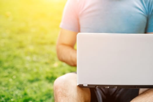 Young man using and typing laptop computer in summer grass. Freelancer working in outdoor park. Sun flare