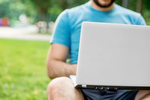 Young man using and typing laptop computer in summer grass. Freelancer working in outdoor park