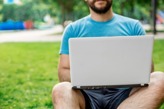 Young man using and typing laptop computer in summer grass. Freelancer working in outdoor park