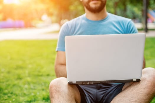 Young man using and typing laptop computer in summer grass. Freelancer working in outdoor park. Sun flare