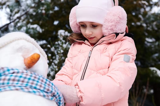 Closeup portrait Caucasian beautiful lovely child girl, wearing pink down jacket and fluffy earmuffs, playing with snowman in snowy winter park, putting around his neck a blue scarf. Happy childhood