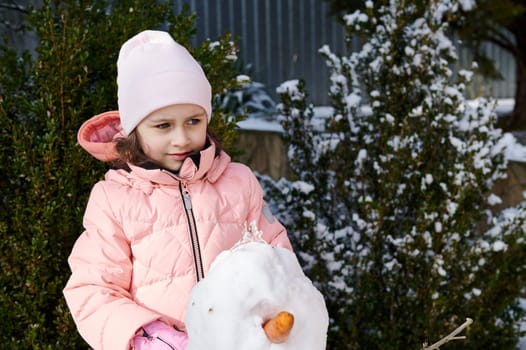 Caucasian cute child girl in warm winter clothes, dreamily looking aside, standing with a snowman in the snow covered backyard. Happy childhood. Beautiful kids. Outdoors winter entertainment for kids