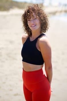 Positive young female athlete with curly hair in top and leggings standing on sandy beach with hands behind back and looking at camera