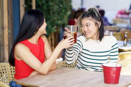 Delighted young Asian female friends, in casual outfits smiling and looking at each other while clinking glasses of drinks at wooden table in cafe during weekend together