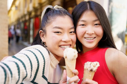 Two young Chinese girls taking a selfie while eating an ice cream cone in the typical streets of Granada, Spain. Concept of Asian people traveling in Europe.