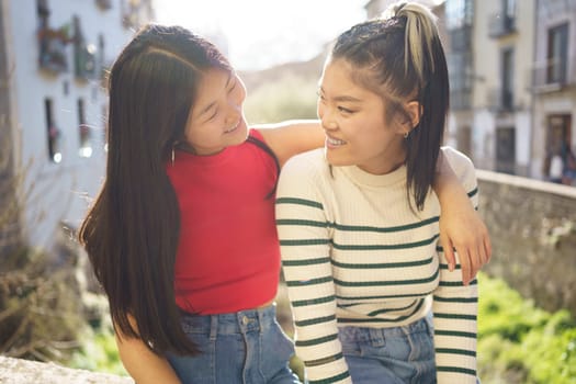 Happy Asian woman sitting with hand on shoulder of friend while looking at each other against blurred background in Granada