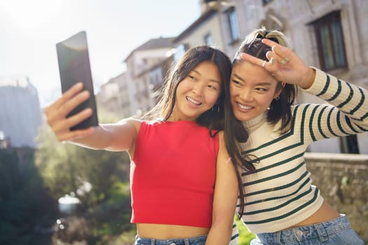 Optimistic Asian girlfriends smiling brightly and showing rock gesture while taking selfie against city buildings in Granada