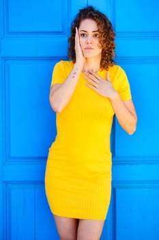 Astonished young female in yellow dress with curly brown hair keeping hand on chest and touching face while looking away against blue wall on street