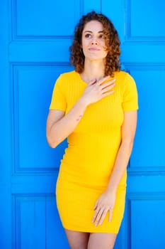Dreamy young female in yellow dress with curly brown hair keeping hand on chest and looking up against blue wall on street