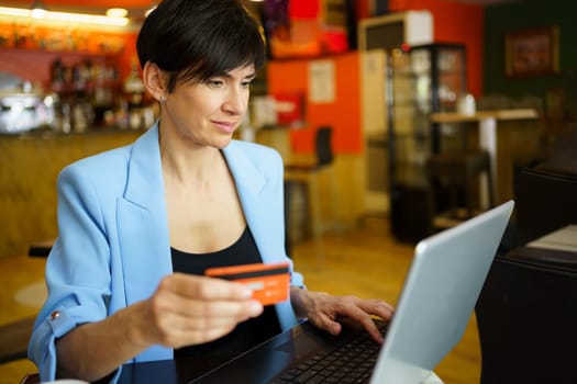 Focused female entrepreneur in smart casual outfit making purchase online using credit card and laptop while browsing in cafe indoors