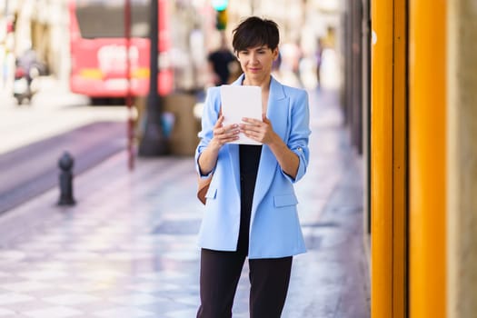 Concentrated female entrepreneur in formal clothes, reading on tablet while standing on city street and looking at screen against blurred background of people and bus in daylight