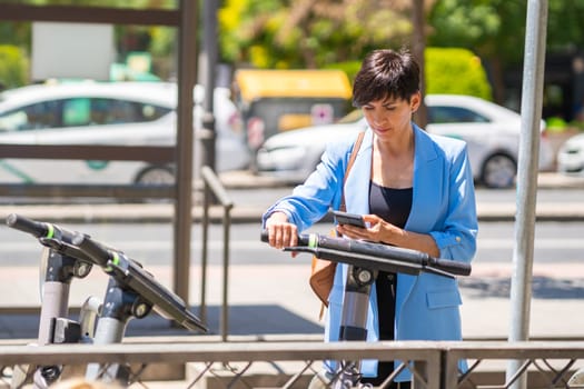 Concentrated adult female in blue jacket scanning QR code on electric scooter with smartphone while standing on sunny street and preparing for riding