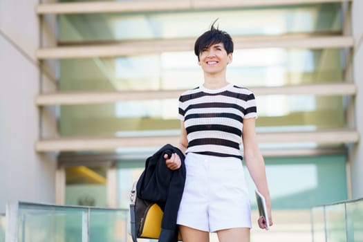 Smiling adult female in black striped shirt and short dress holding jacket and tablet while standing on balcony of modern building with blurred background