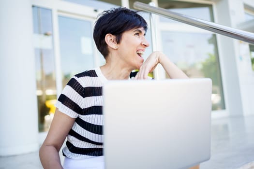 Positive adult female freelancer with short hair smiling and looking away while sitting on paved stairs and working remotely on laptop