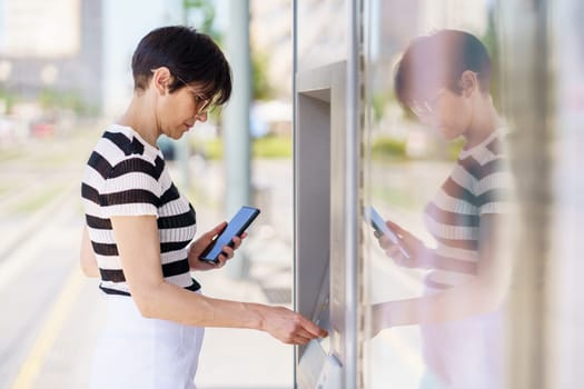 Side view of focused adult female in casual outfit and eyeglasses standing with smartphone and buying ticket in modern terminal during trip