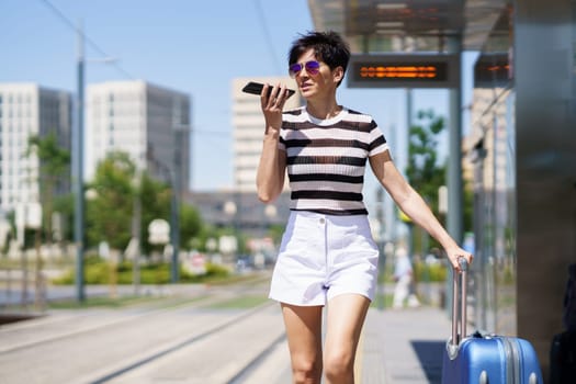 Positive young female in trendy outfit, and sunglasses with suitcase standing on city street by railroad and recording audio message on mobile phone in bright daylight
