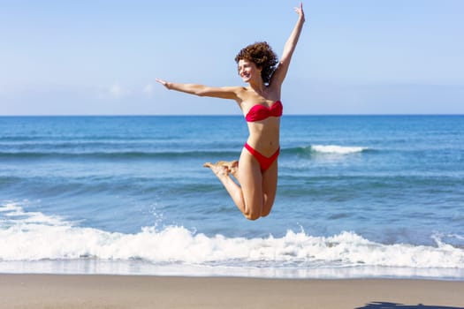 Excited young female in pink bikini stretching arms and jumping in air with folded legs up, while smiling and looking away in daylight against rippling sea and blue sky