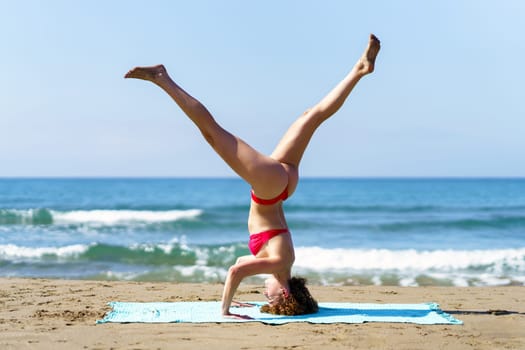 Side view of calm female in bikini performing Tripod Headstand while having session alone on coast of ocean