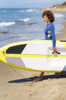 Side view of young female surfer in wetsuit on sandy beach with SUP board at sandy shore with wavy water, while walking and looking at sea in sunny daylight
