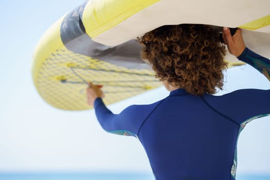 Back view of unrecognizable curly haired female in blue swimwear lifting white yellow SUP board on head while facing blue sea in bright daylight