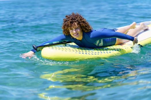 Glad curly woman in wetsuit looking at camera while lying and pulling paddleboard through shining surface of sea in sunshine