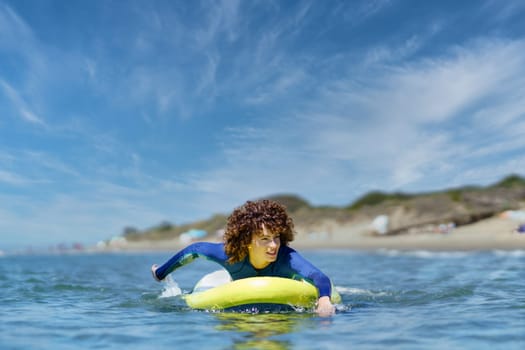 Young curly haired woman in wetsuit lying on standup SUP board paddling with hands in sea against blue sky on sunny summer day