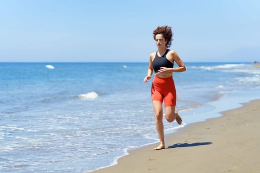 Concentrated barefoot young female in sportswear and with curly hair looking away, while jogging in daylight on sandy beach near foamy seawater against blurred blue sky