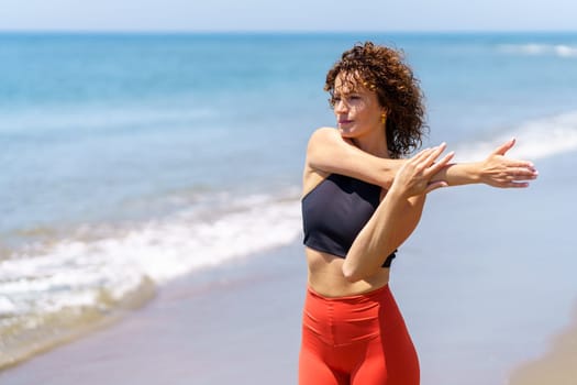 Pensive young sportswoman with curly hair in sportswear stretching arm while standing near waving sea and looking away