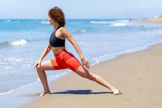 Full body side view of curly haired female athlete in activewear doing lunges near waving sea on sunny day