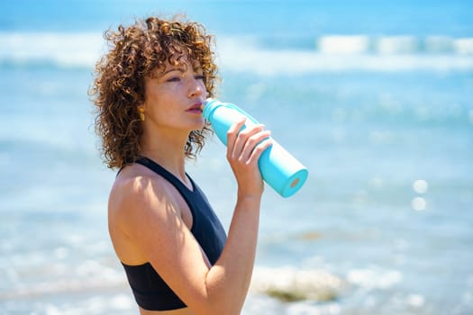 Side view of young female athlete with curly hair standing on seashore and drinking water from blue plastic bottle after workout