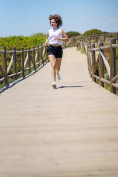 Full body of fit female athlete in sportswear running on wooden path and looking away during sunny summer day