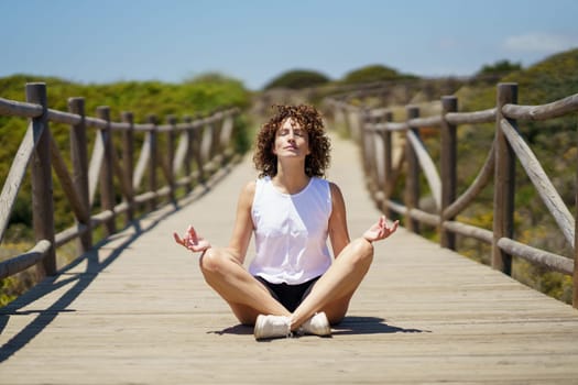 Young curly haired female in casual clothes sitting on wooden walkway and meditating, with closed eyes while doing lotus pose against blurred green hill under blue sky