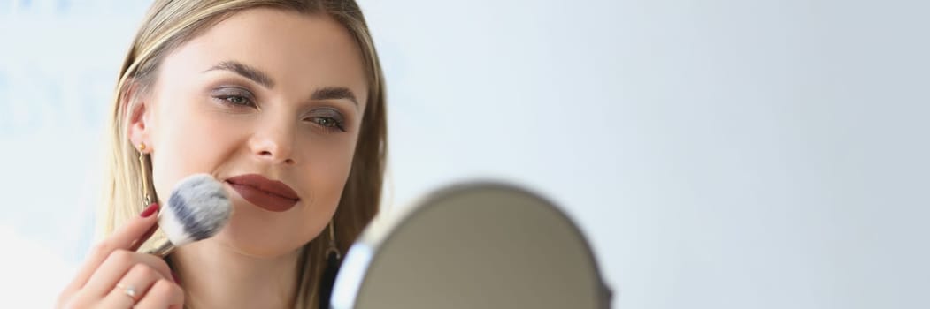 Smiling young woman looks in the mirror and paints herself with blush brushes. Makeup artist girl doing makeup learning with beauty and cosmetics video tutorials