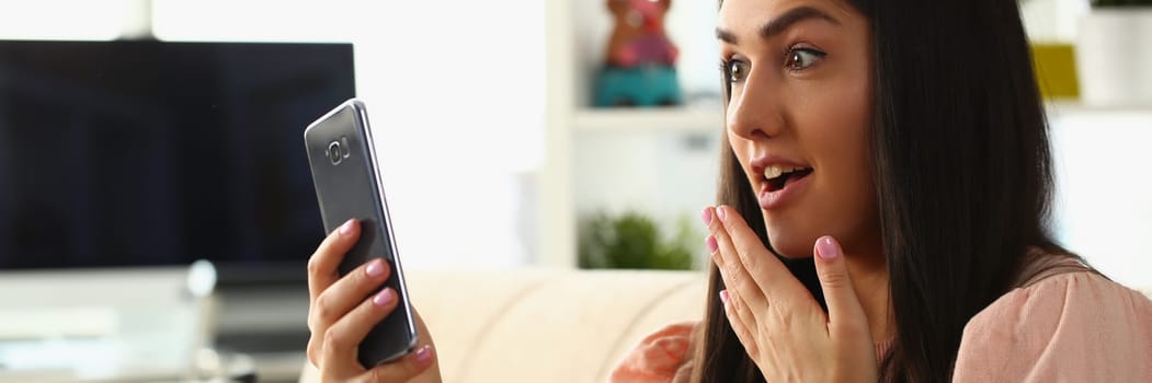 Woman surprised in shock looks at smartphone screen while sitting on couch at home. Emotion joy delight and good news concept